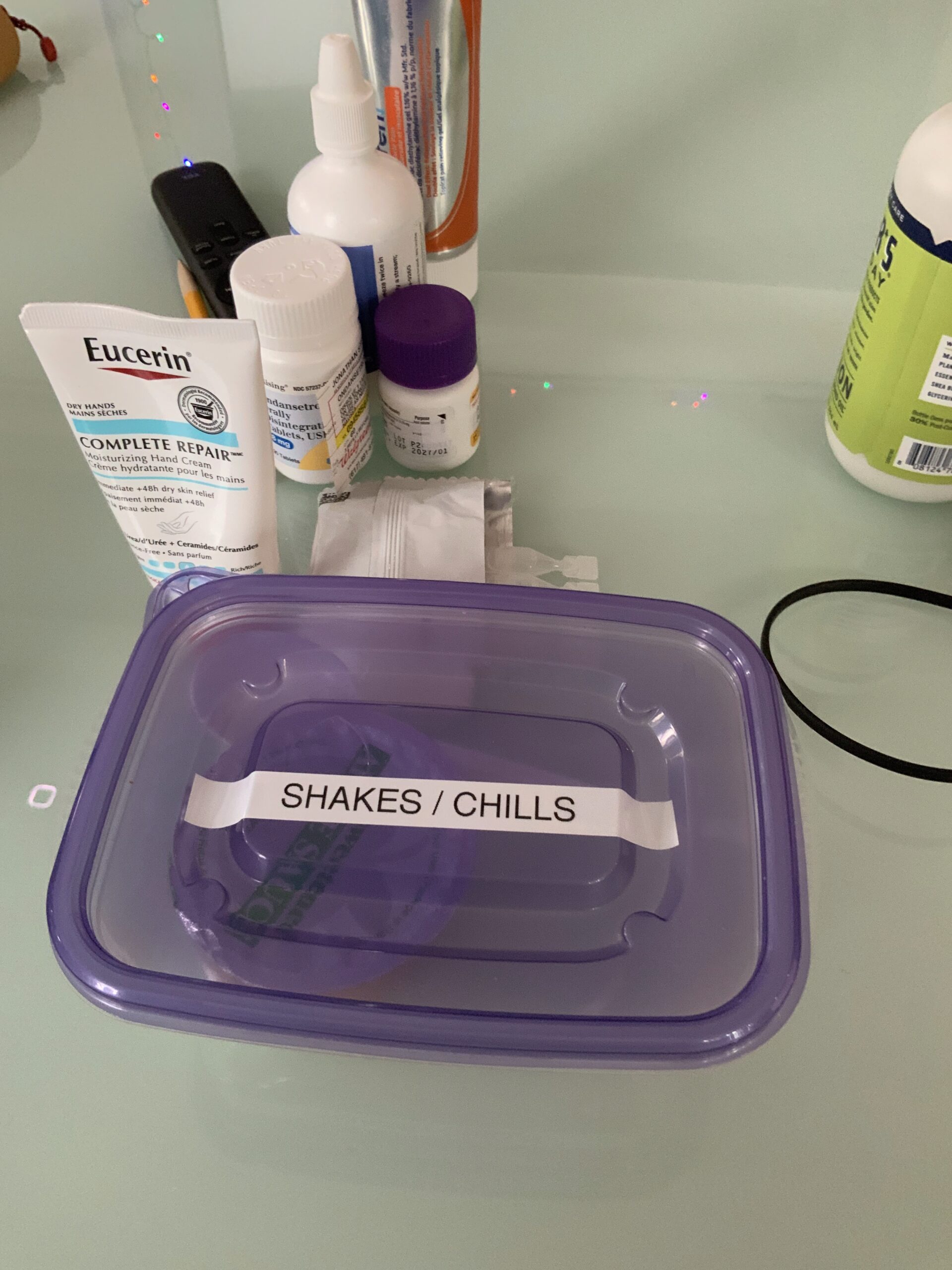 A plastic container labelled "shakes/chills" in all caps on its purple lid. Behind it is some hand cream, nasal spray, and some other pill containers.