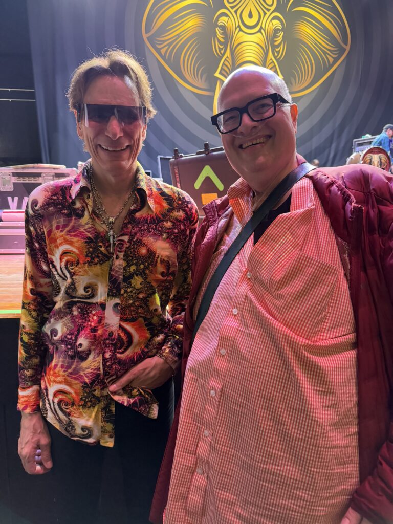 Jonathan in an orange shirt and nasal cannula, to the right of Steve Vai, in a multicoloured shirt and sunglasses. 