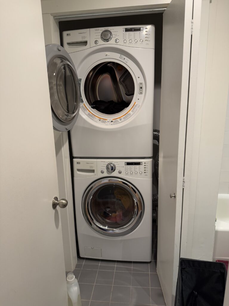 A front-loading dryer on top of a front loading washer, wedged into the corner of a bathroom. There are two doors making the space even tighter.